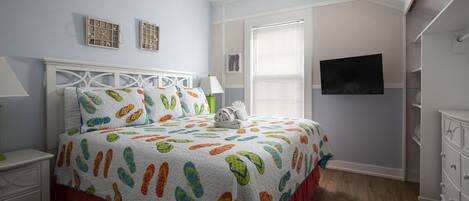 A bedroom with a bed, nightstand, dresser, and a TV on the wall.
