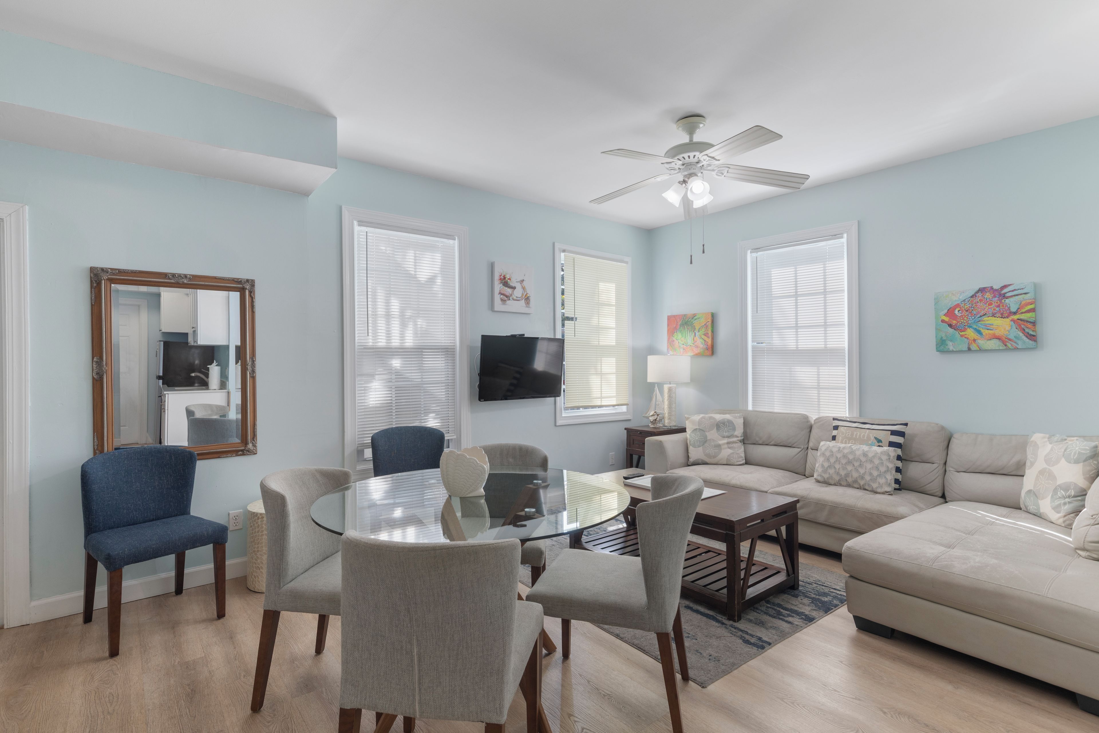 A living room with a round glass table, two blue chairs, a beige sofa, and a ceiling fan.