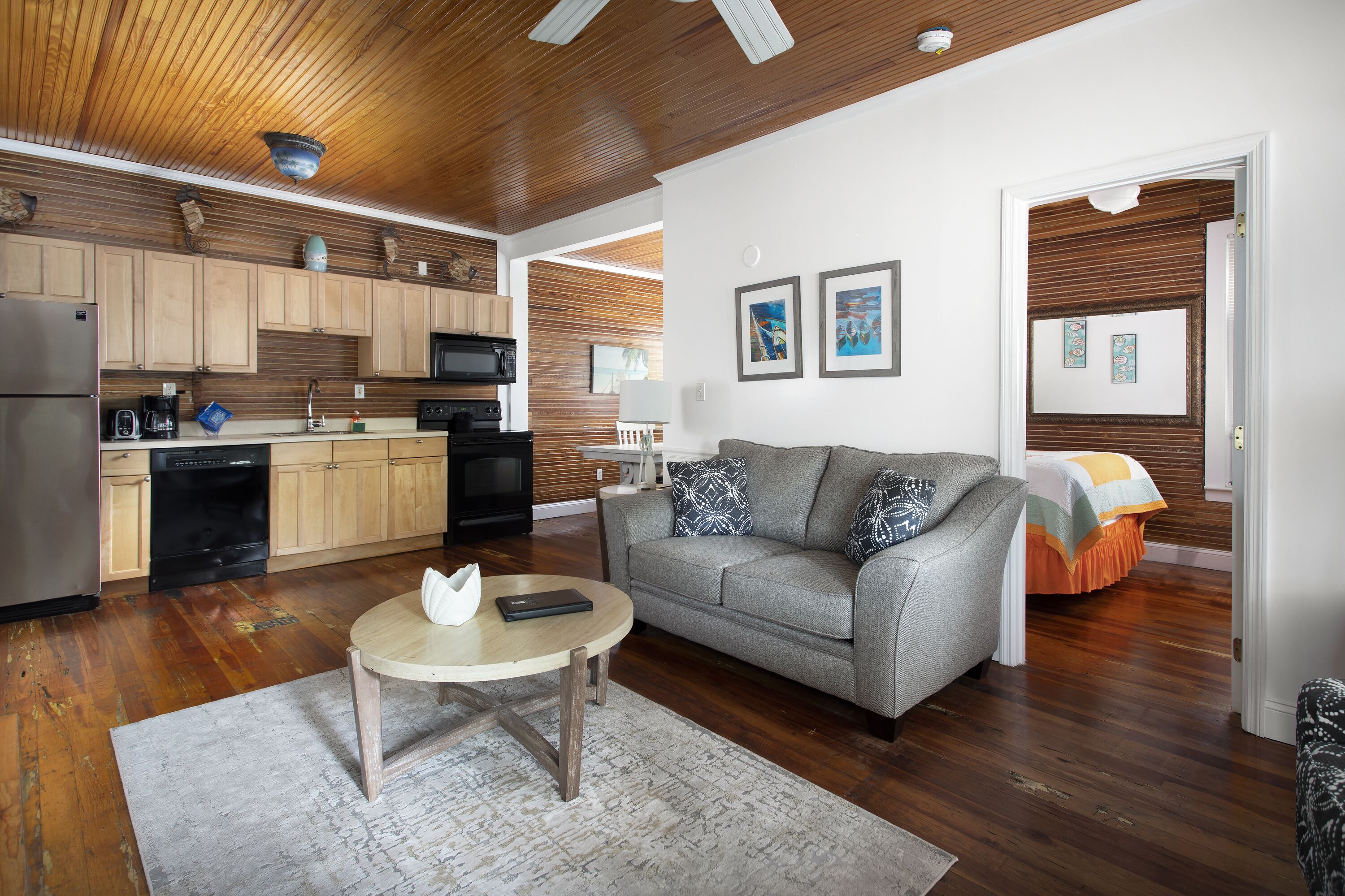A modern kitchen with wooden cabinets, a dining area with a round table, and a living room with a sofa and a bed in the background.