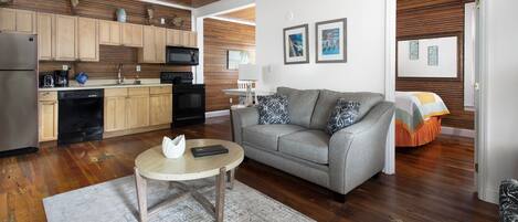 A modern kitchen with wooden cabinets, a dining area with a round table, and a living room with a sofa and a bed in the background.