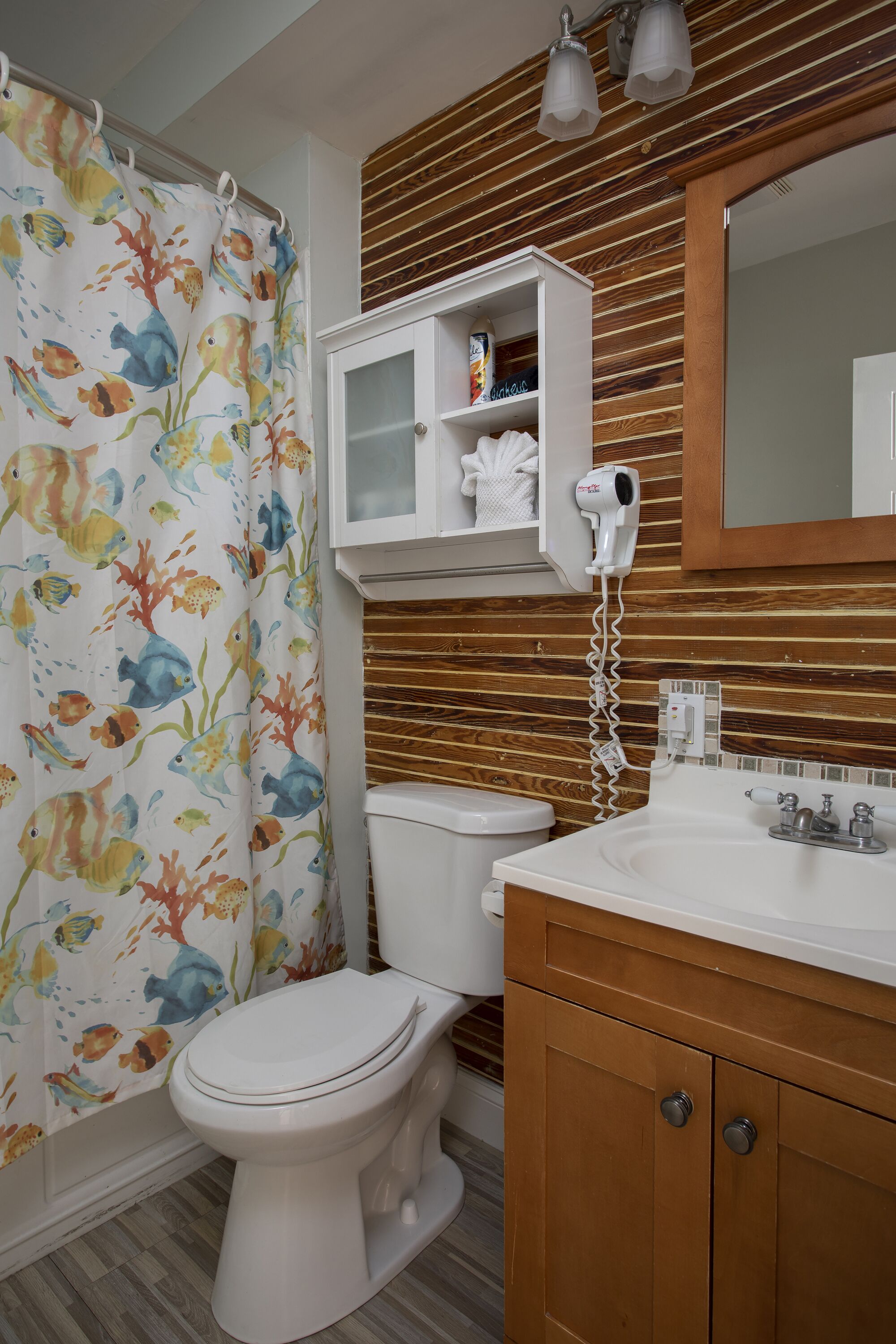 A bathroom with a shower curtain featuring colorful fish, a white toilet, a wooden vanity with a mirror, and a hairdryer mounted on the wall.