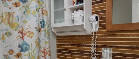 A bathroom with a shower curtain featuring colorful fish, a white toilet, a wooden vanity with a mirror, and a hairdryer mounted on the wall.