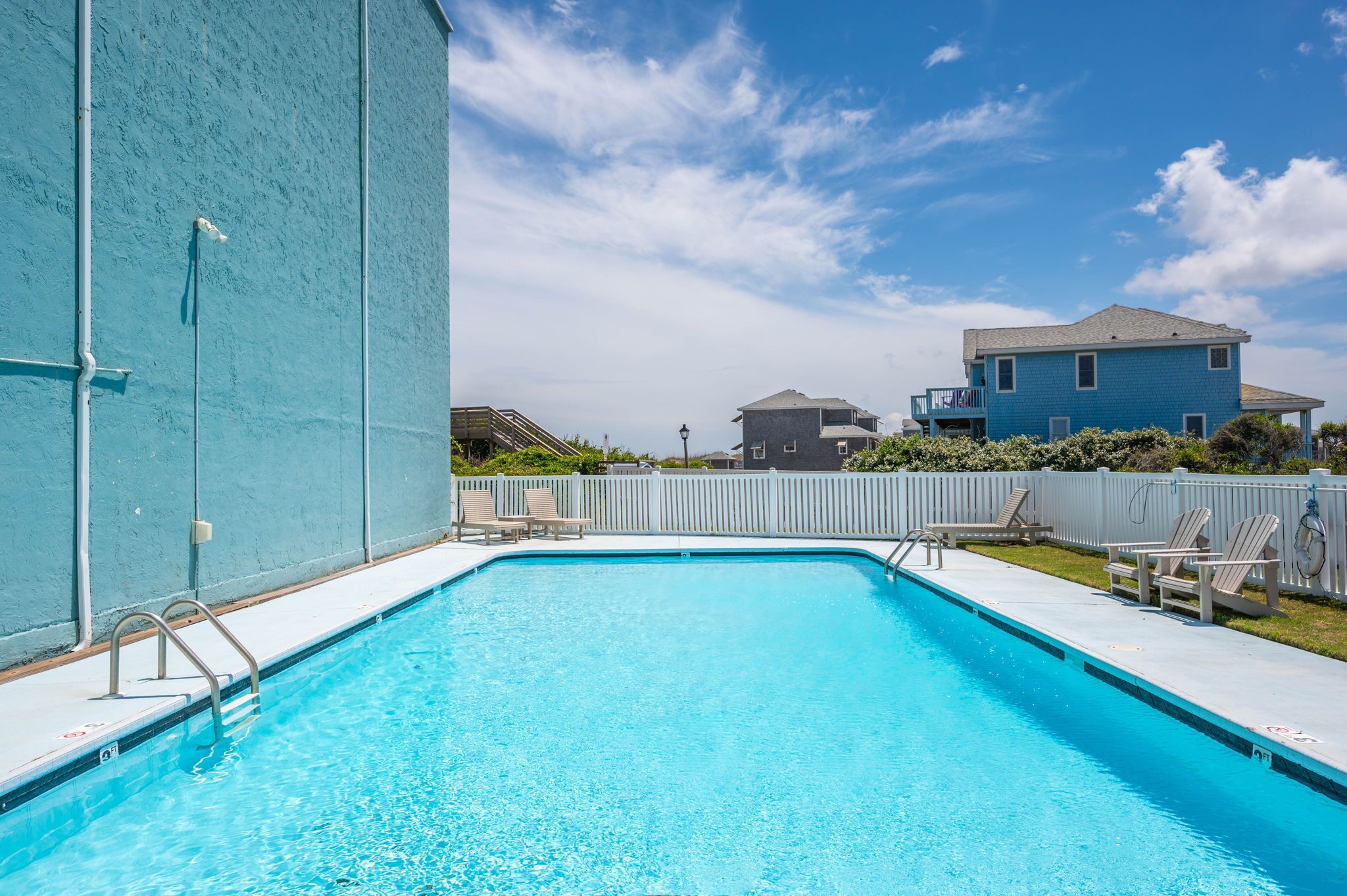 A rectangular swimming pool with a metal ladder on one side, surrounded by a white fence. There are lounge chairs and a lifebuoy on the fence. In the background, there is a blue house with a grey roof.