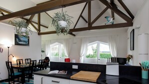 Una cocina moderna con armarios blancos, encimeras negras y techo de madera. Hay dos plantas colgantes, un comedor con sillas y una mesa, y vistas al verdor exterior.