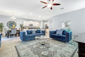 A modern living room with two blue sofas, a glass coffee table, and a dining area with a large clock and cabinets.
