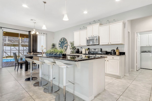 A modern kitchen with white cabinets, a central island with bar stools, stainless steel appliances, and a large clock on the wall.