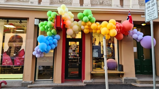Un bâtiment avec une arche de ballons colorés à l’entrée, un panneau indiquant un parking et une devanture de magasin avec une vitrine.
