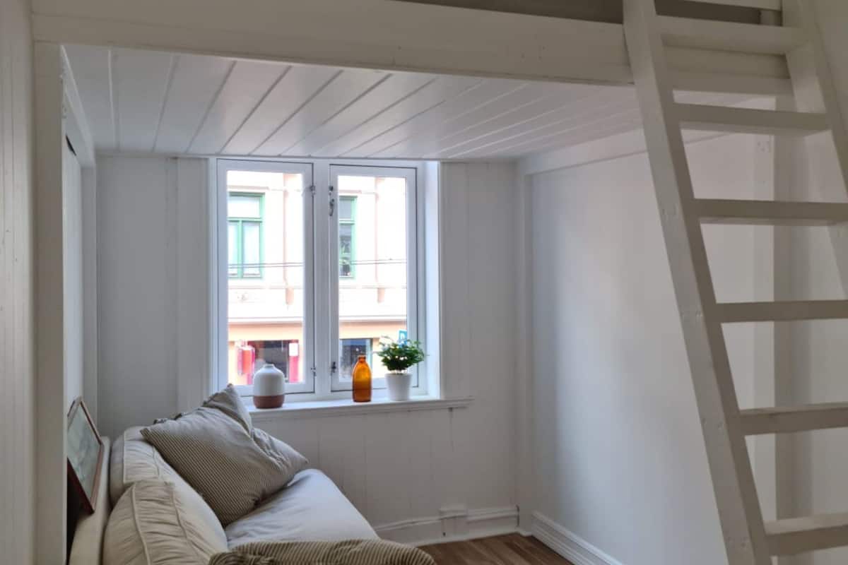 A small, cozy room with a loft bed, a window with a view of buildings, a ladder, and a table.