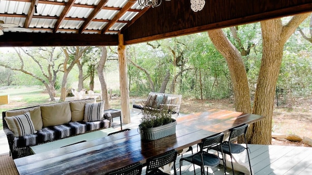 An outdoor patio with a wooden table, chairs, a sofa, and a ceiling fan. - Farmhouse: Sleeps 6 | Soaking Tub | Fire Pit | Wimberley TX (Wimberley)