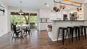 A modern kitchen with white cabinets, a wooden dining table, and black bar stools. - Farmhouse: Sleeps 6 | Soaking Tub | Fire Pit | Wimberley TX (Wimberley)