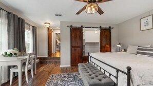 A bedroom with a bed, a desk, a chair, a bench, a ceiling fan, and a door. - Farmhouse: Sleeps 6 | Soaking Tub | Fire Pit | Wimberley TX (Wimberley)