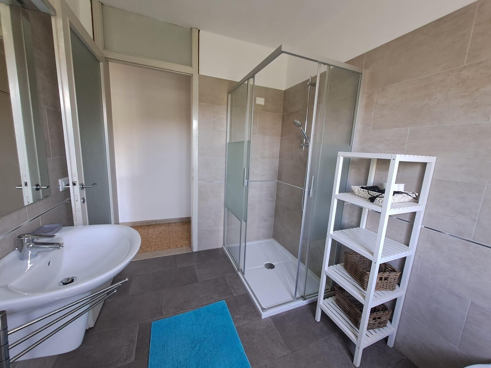 Une salle de bain avec un lavabo blanc, une cabine de douche en verre et une étagère blanche avec des paniers.