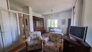 A living room with two floral armchairs, a TV, a cabinet, a chandelier, and a dining area with a table and chairs.