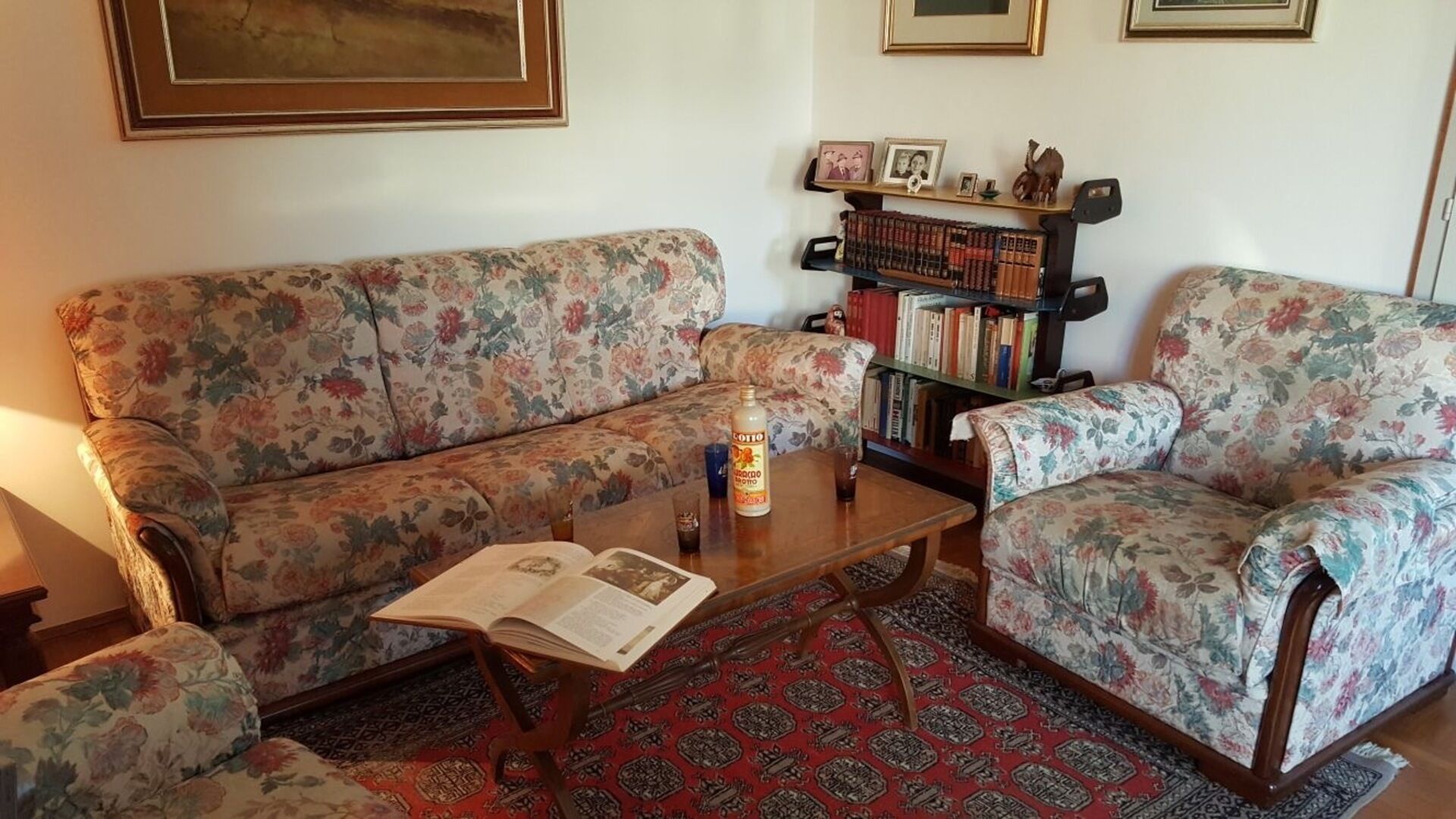 Ein Wohnzimmer mit einem blumigen Sofa, einem Holztisch mit einem Buch und einer Flasche, einem Bücherregal mit Büchern und gerahmten Bildern sowie einem gemusterten Teppich.