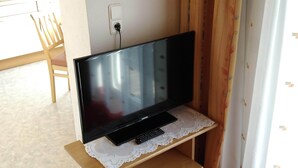 Televisió sobre un prestatge de fusta amb el comandament a mà, una presa de corrent a la paret, una cadira i una catifa amb dibuixos.