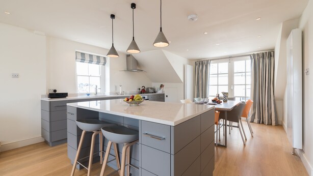 A modern kitchen with a central island, bar stools, pendant lighting, and a dining area with a table and chairs. - Dawson Place (London)