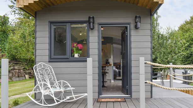 Une petite cabane en bois dotée d’une terrasse, d’une chaise berçante et d’une porte donnant sur un intérieur comprenant une cuisine et un lit.