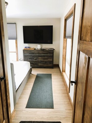 A bedroom with a bed, a dresser, a flat-screen TV mounted on the wall, and a wooden floor.