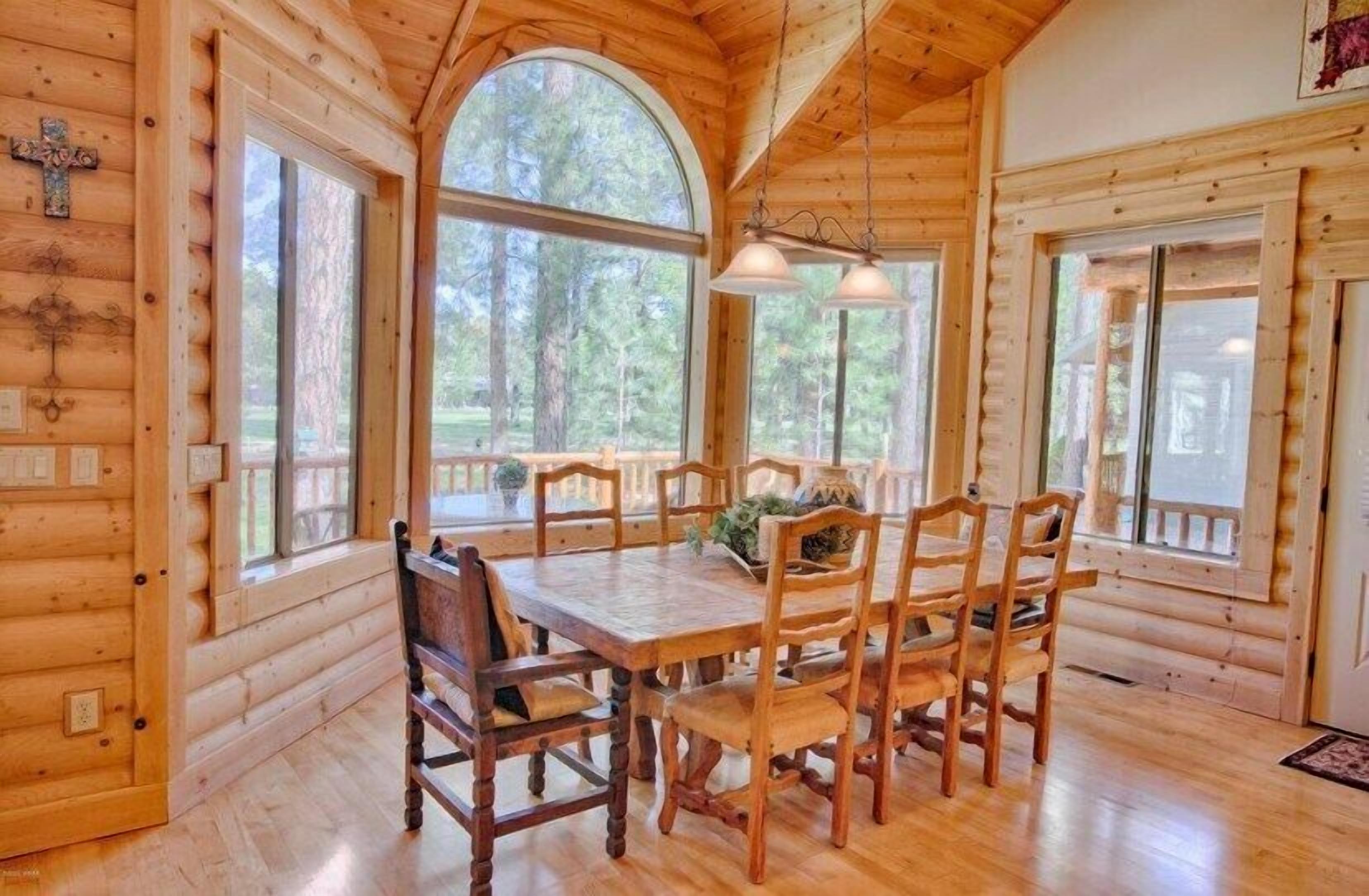 A wooden cabin interior with a large dining table, multiple chairs, and large windows offering views of the surrounding forest.