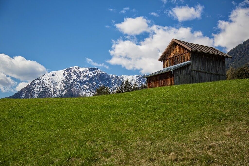 Una cabaña de madera con techo inclinado sobre una colina cubierta de hierba, con vistas a una montaña nevada bajo un cielo despejado.