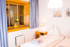 A neatly made bed with white linens, a bedside table with a lamp, a window with curtains, and a wall clock.