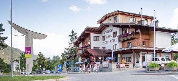 A hotel with a ski shop and kiosk on the ground floor, a mountain view, and a pedestrian area with people and cars.
