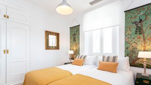 Quarto de hotel com duas camas, paredes brancas, uma janela grande e obras de arte nas paredes.