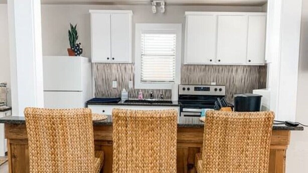 Een keuken met drie rieten stoelen, witte kastjes, een fornuis en een raam met zonwering.