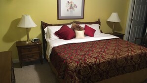 Ein ordentlich bezogenes Bett mit gemustertem Überwurf, zwei Nachttischlampen, ein gerahmtes Blumenbild an der Wand und eine Tür auf der rechten Seite.