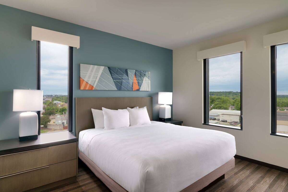 A hotel room with a large bed, two bedside tables with lamps, a view of the city through large windows, and a modern abstract painting on the wall.