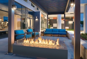 Un área de estar al aire libre con un hogar, sillas acolchadas azules y un edificio moderno con grandes ventanales al fondo.