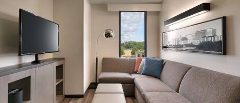 A modern living room with a flat-screen TV, a sectional sofa, a coffee table, and a large window offering a city view.