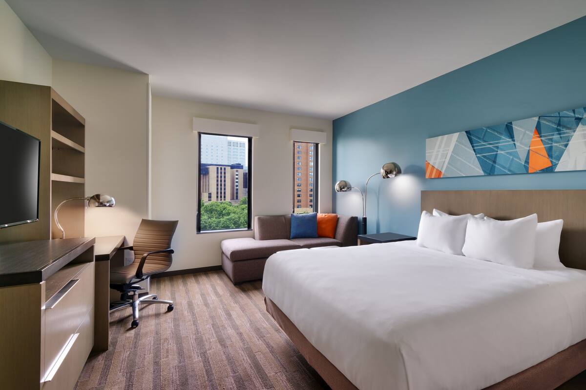 A hotel room with a large bed, a desk, a chair, a TV, and a window with a city view.