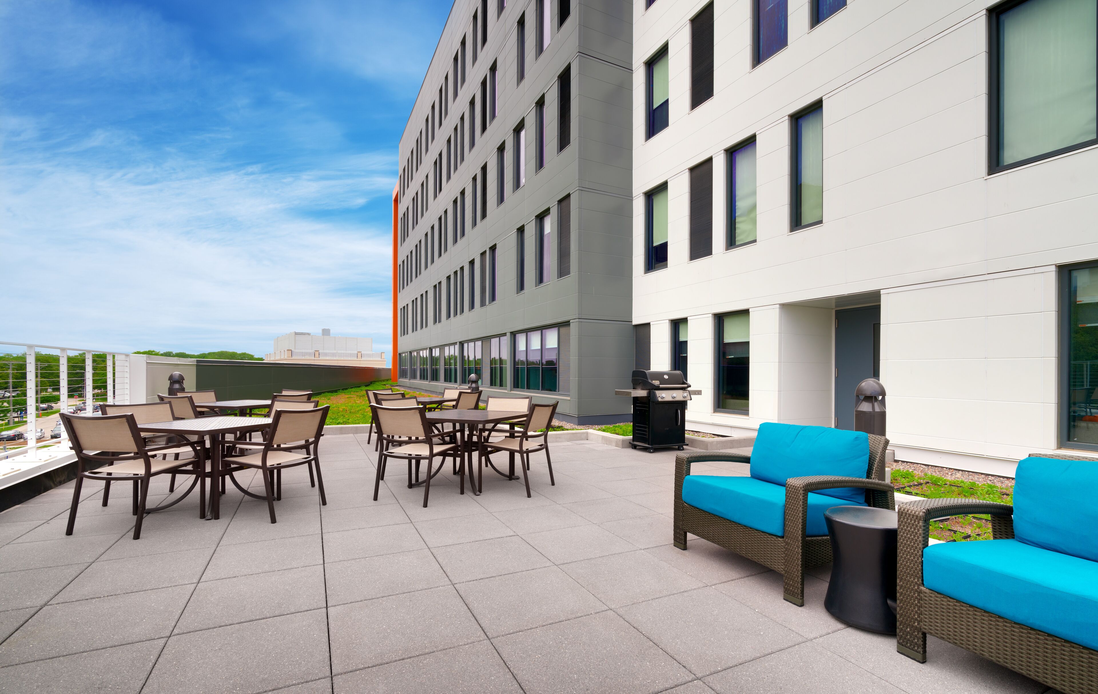 A rooftop patio with outdoor furniture, a grill, and a view of a modern building.