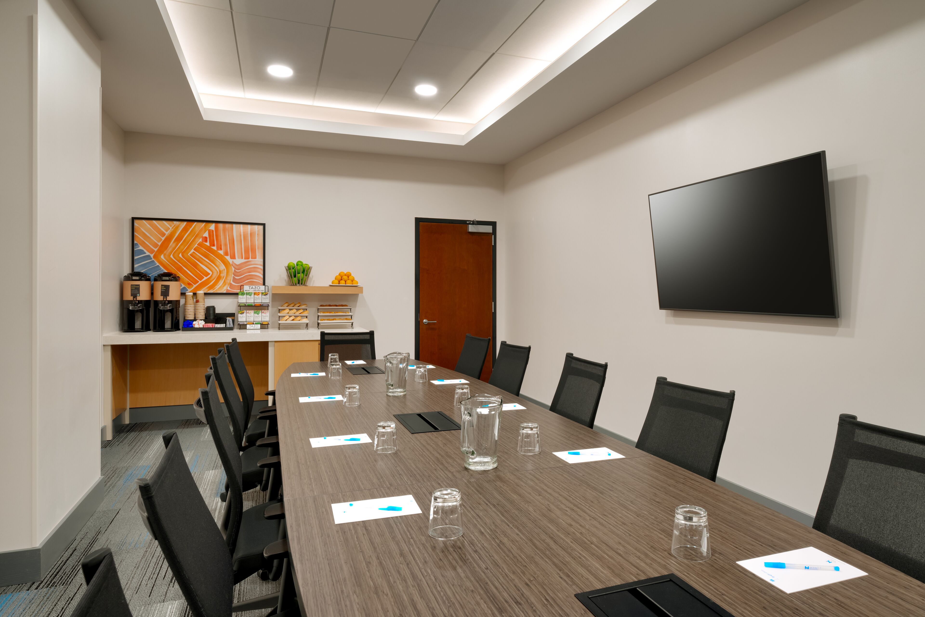 A conference room with a long table, chairs, a TV on the wall, and a door.