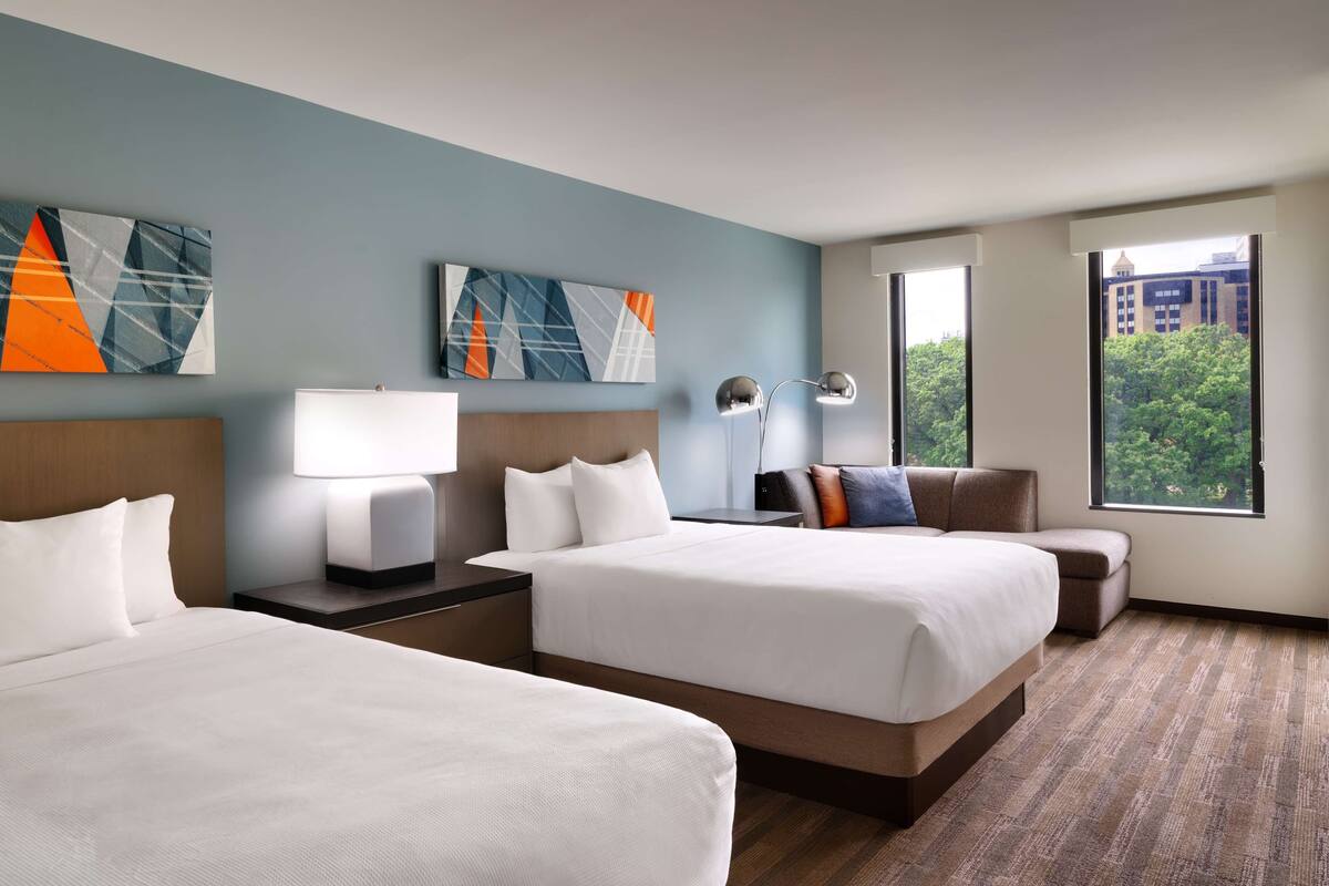 A hotel room with two beds, a desk, a chair, and a large window offering a city view.