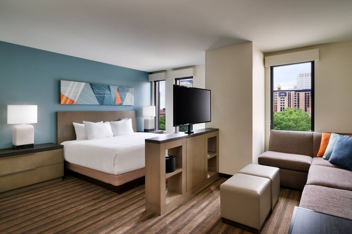 A hotel room with a bed, a desk with a TV, a sofa, and a window with a city view.