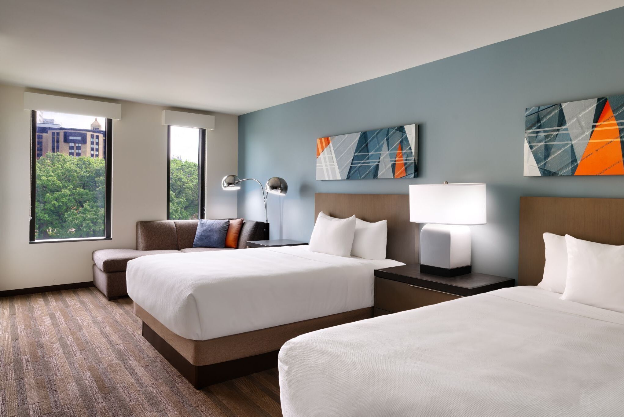 Ein Hotelzimmer mit zwei Betten, einer Sitzecke, einem Schreibtisch und einem großen Fenster mit Blick auf die Stadt.