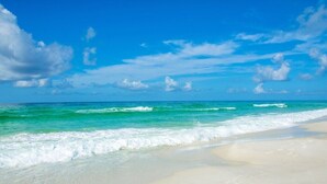 Une plage à l’eau turquoise et limpide, bordée de sable blanc, sous un ciel d’un bleu éclatant.