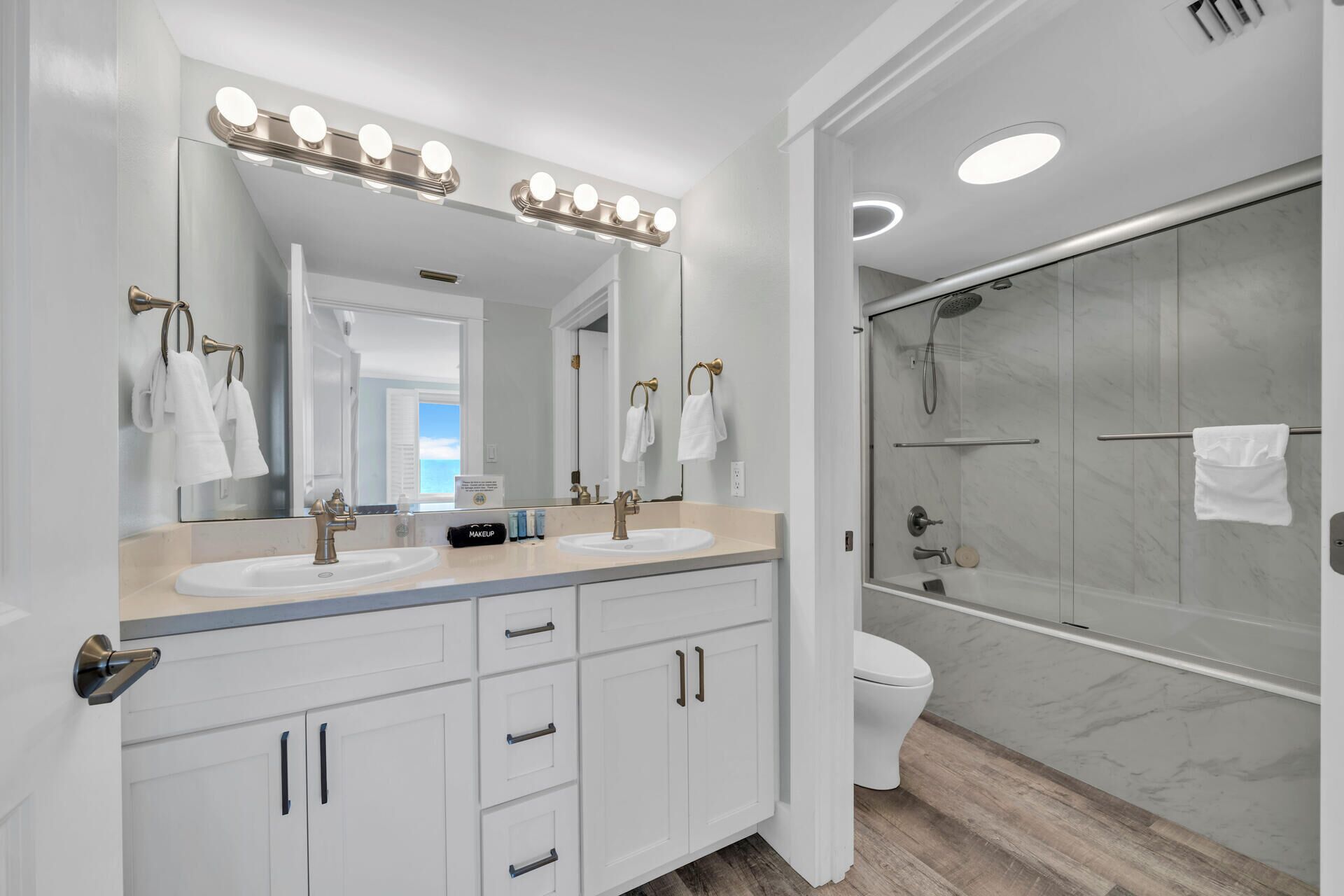 A modern bathroom with two sinks, a large mirror with lights, a shower with glass doors, and a toilet.