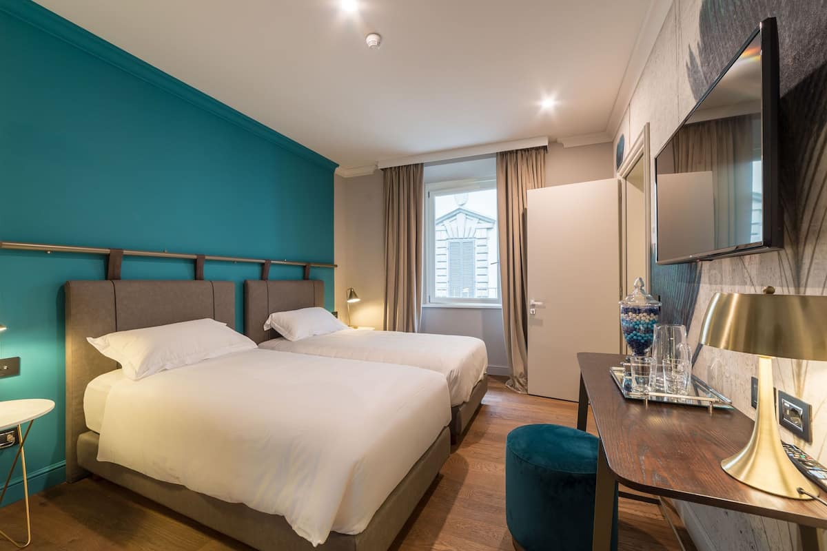 Habitación de hotel con dos camas, un escritorio con lámpara, un televisor y una ventana con cortinas.
