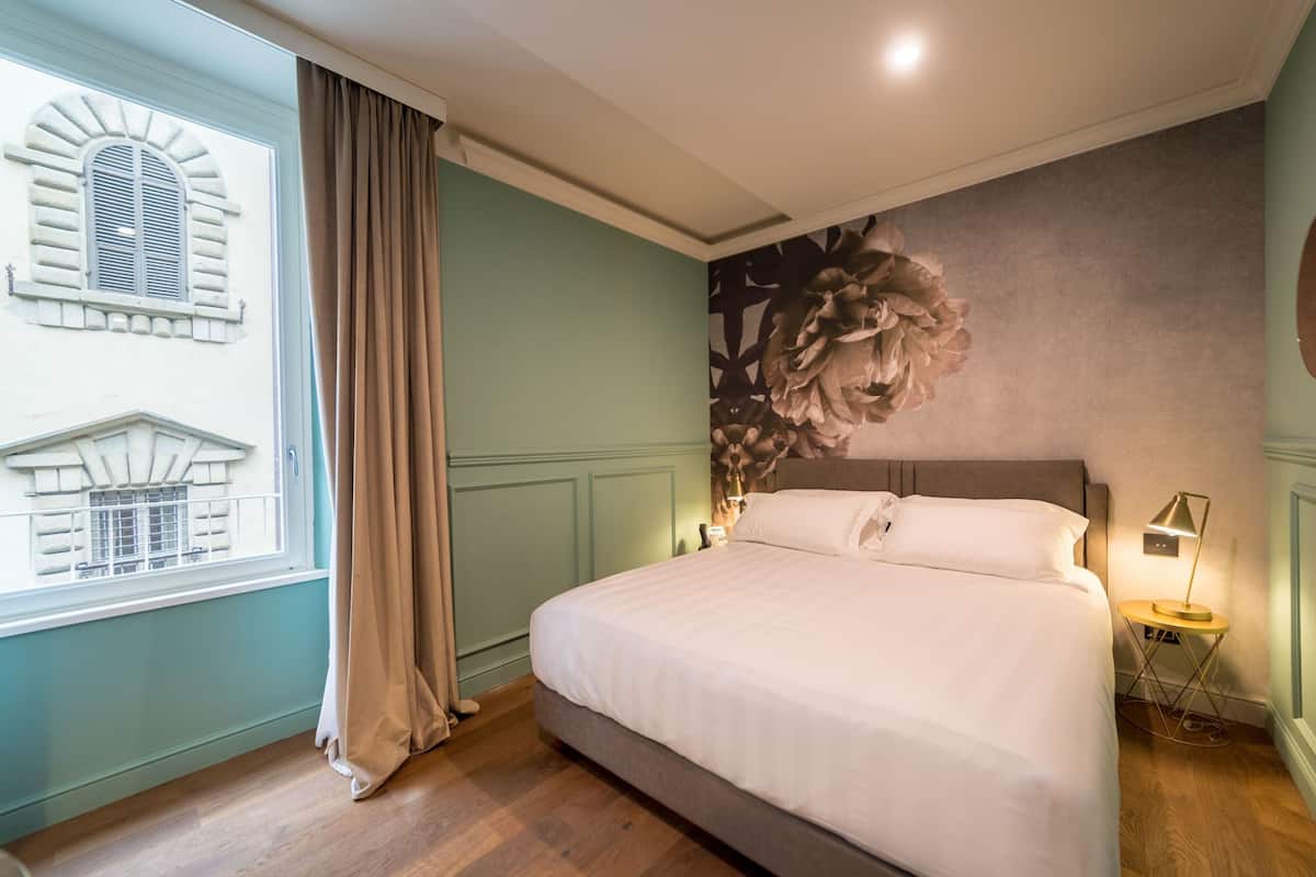 Una cama bien hecha en una habitación con un ventanal grande, un mural en la pared y una mesita de noche con una lámpara.