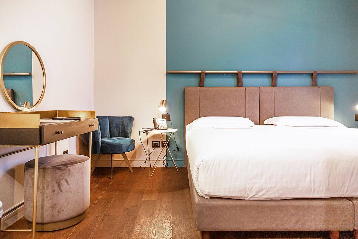 Una habitación de hotel con una cama, un escritorio con una silla, un reposapiés redondo, una mesita de noche con una lámpara y un espejo.