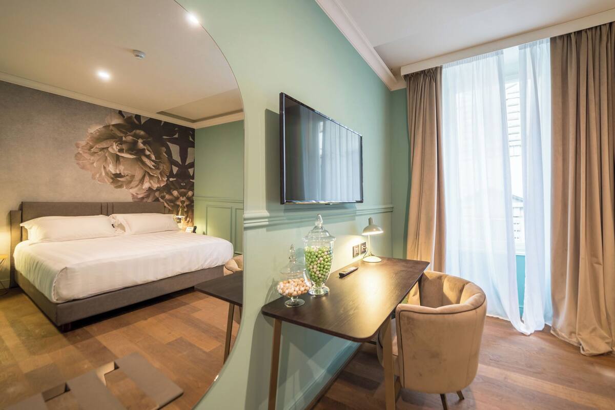 Habitación de hotel con cama, escritorio, silla, televisor y ventana con cortinas.