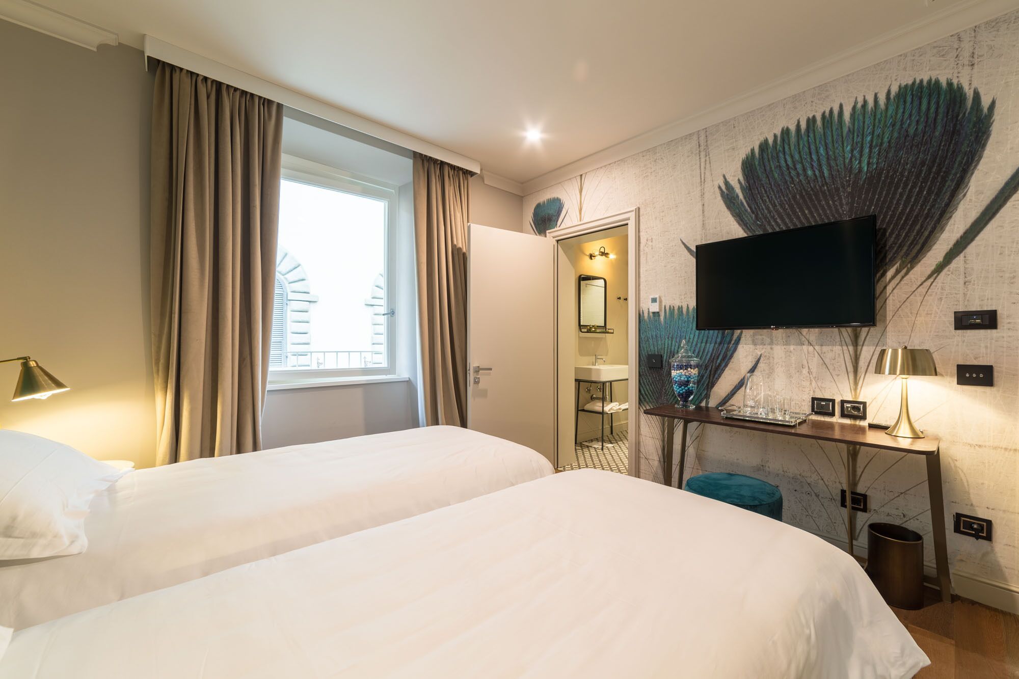 A hotel room with two beds, a television, a desk with a lamp, and a peacock feather mural on the wall.