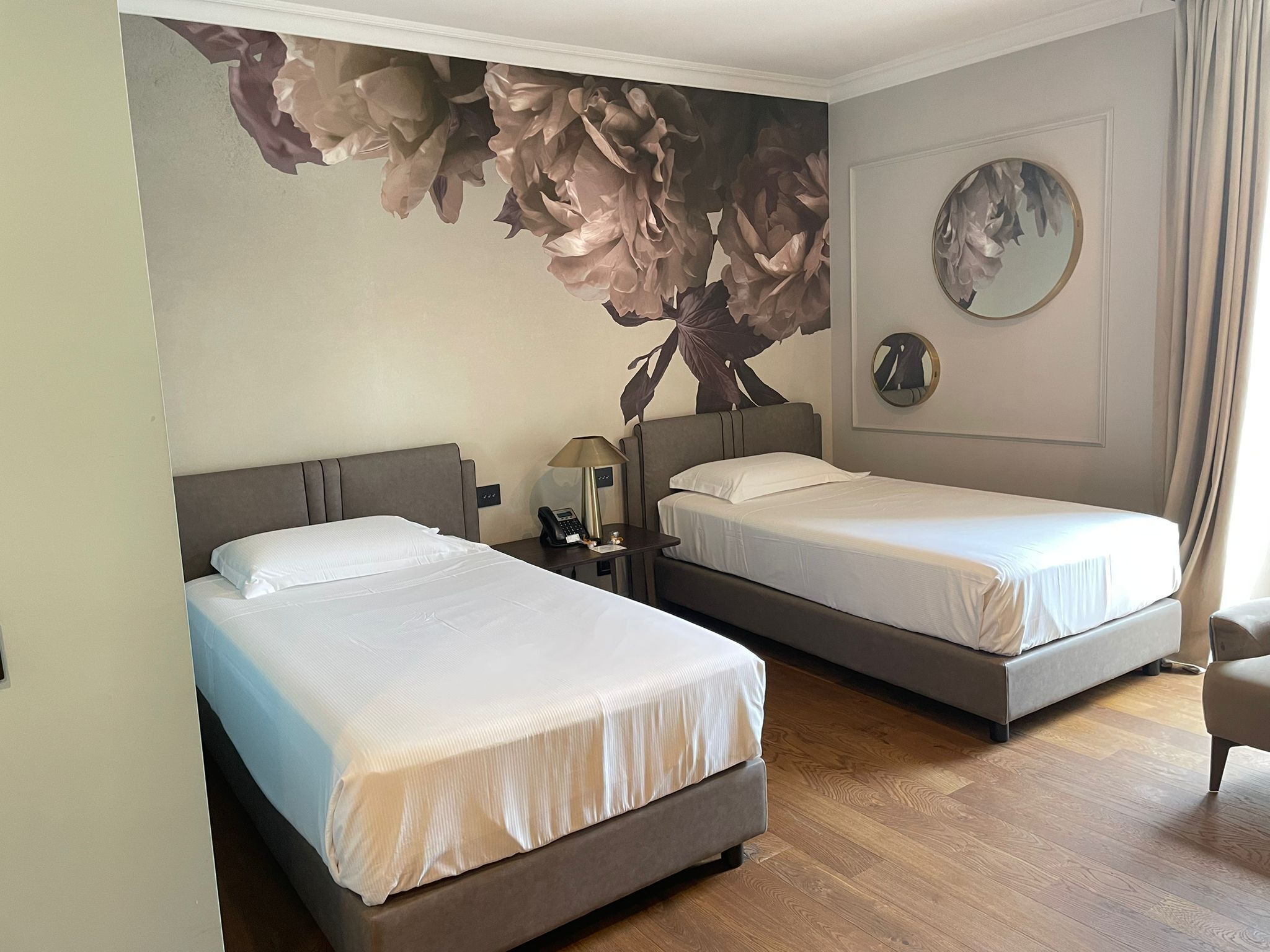 Habitación de hotel con dos camas, un mural grande con motivos florales en la pared, un espejo redondo y suelo de madera.