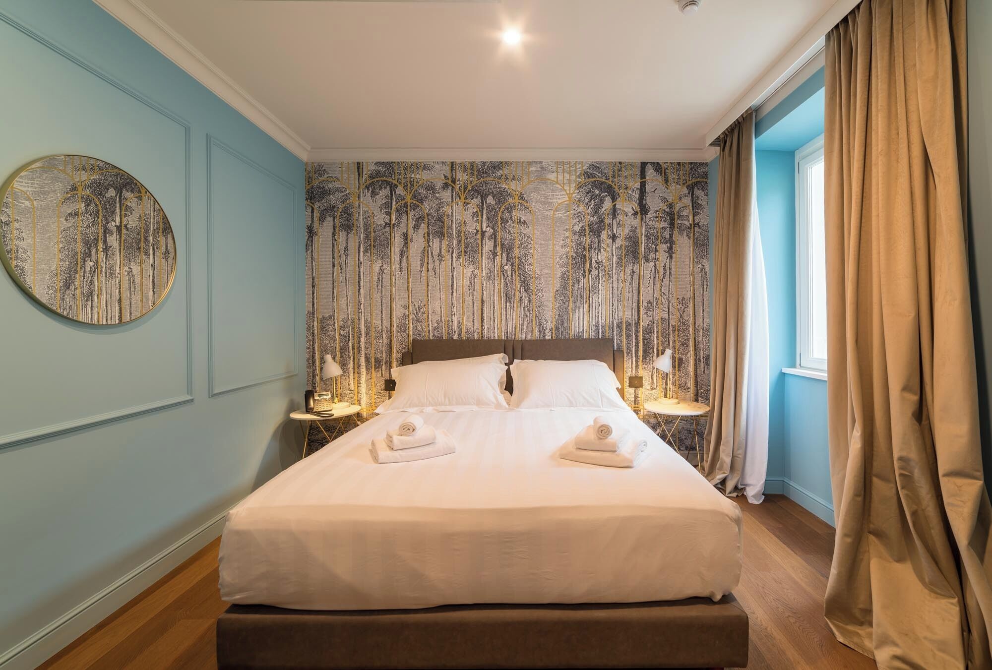 A neatly made bed with two pillows and two towels, a round mirror on the wall, and a patterned tapestry on the headboard.