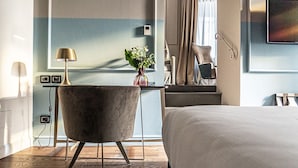 Habitación de hotel con cama, escritorio con lámpara, silla, florero con flores y un ventanal con cortinas.