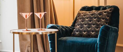 A room with a blue velvet armchair, a round table with two martini glasses, a floor lamp, and a patterned cushion.