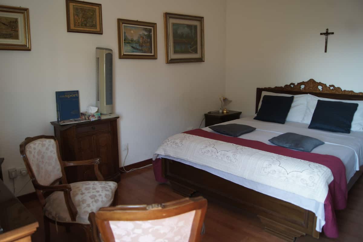 Un dormitorio con cama de madera, dos almohadas, una mesita de noche, una silla, un escritorio con computadora y cuadros en la pared.